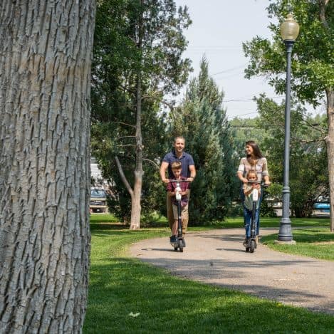 A man and child ride an electric scooter while a woman and another child ride a second scooter on a paved path in a park.