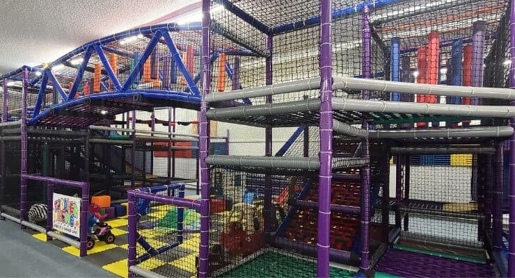 Indoor children's play structure with climbing frames, slides, padded obstacles, and netting in a brightly colored indoor play area.