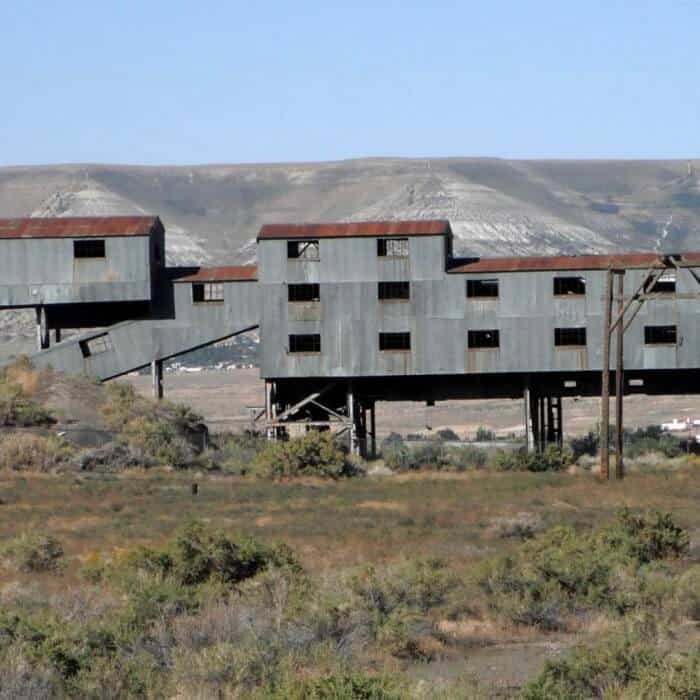 An old, abandoned industrial building with rusted metal walls stands elevated above scrubby vegetation in a rural landscape.