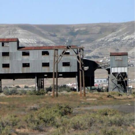 Large, abandoned industrial building with rusted metal siding stands on stilts in a dry, grassy landscape with hills in the background.