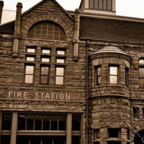 Stone fire station building with arched windows, a tower feature, and "FIRE STATION" sign on the front. Image is in sepia tone.