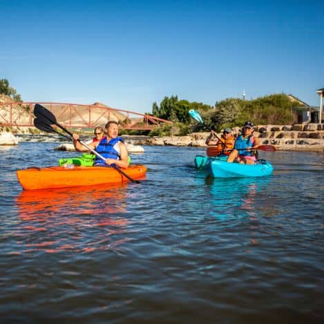 Four people wearing life jackets paddle kayaks on a calm river with a bridge and stone steps in the background under a clear blue sky.