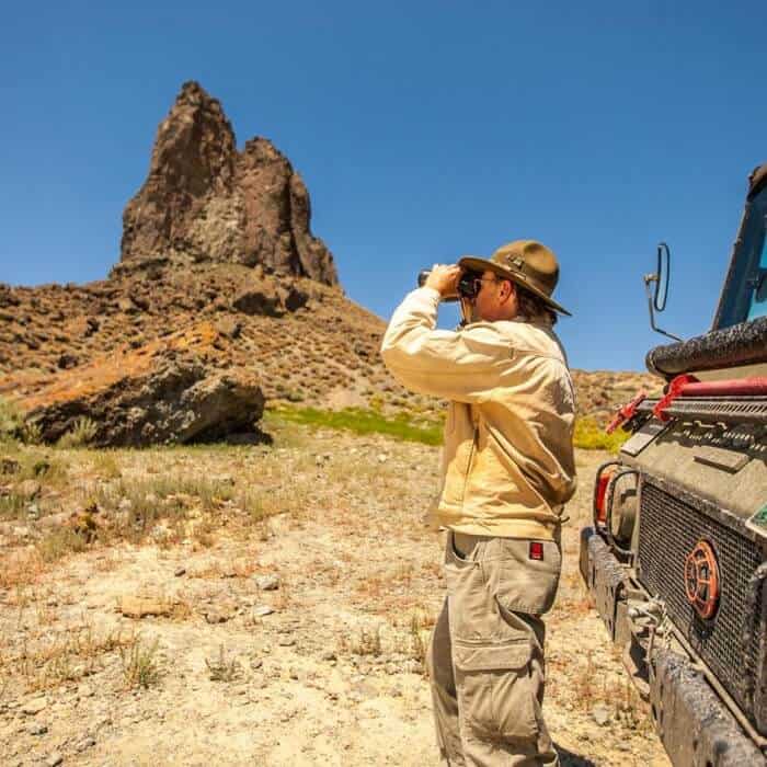 Person in outdoor gear uses binoculars near a rugged rock formation, standing beside an off-road vehicle in a dry, rocky landscape under a clear blue sky.