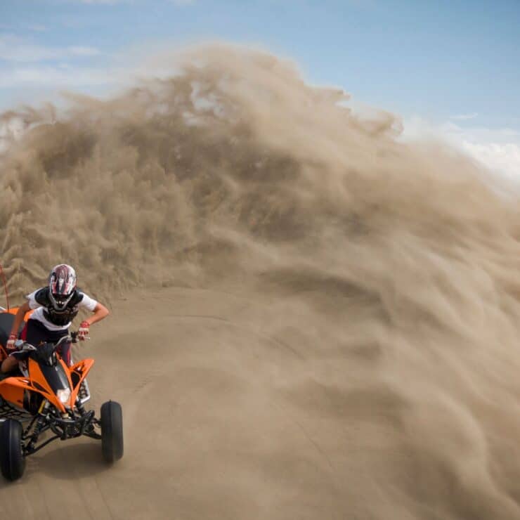Person riding an orange ATV on sand dunes, creating a large spray of sand behind them under a partly cloudy sky.