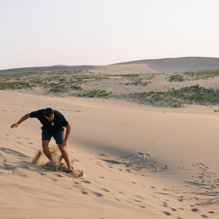 A person wearing a black shirt and shorts climbs up a sandy dune in a desert landscape with sparse vegetation under a clear sky.