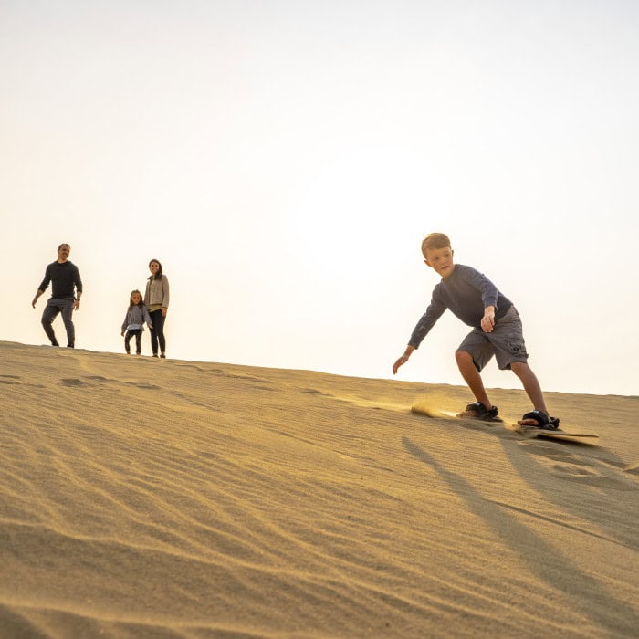 A boy sandboards down a dune as three people stand watching in the background under a bright sky.