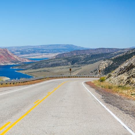 A paved road curves downhill toward a blue lake, surrounded by arid hills and rocky mountains under a clear blue sky.