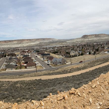 A residential neighborhood sits below rocky hills under a cloudy sky, with houses, roads, and sparse vegetation visible in the landscape.