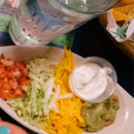 Oval dish with shredded lettuce, diced tomatoes, shredded cheese, guacamole, and a cup of sour cream next to a glass of water and a basket of tortilla chips.