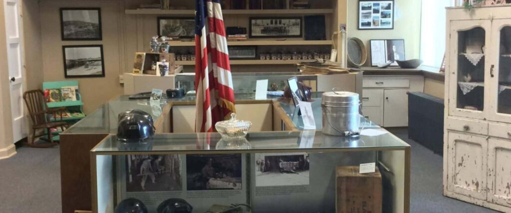 Small museum room with historical military artifacts in glass cases, an American flag, old photos, helmets, and vintage household items displayed around the room.