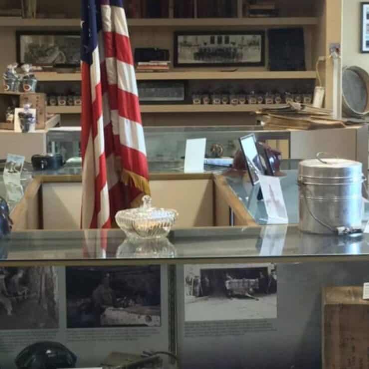 A museum exhibit features glass display cases with historical artifacts, photographs, helmets, an American flag, and framed pictures on the walls.