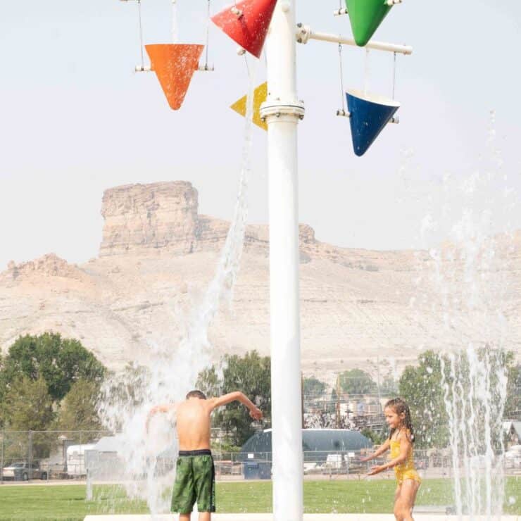 Two children play under a splash pad with tipping buckets and jets of water, with a rocky landscape and sparse trees in the background.