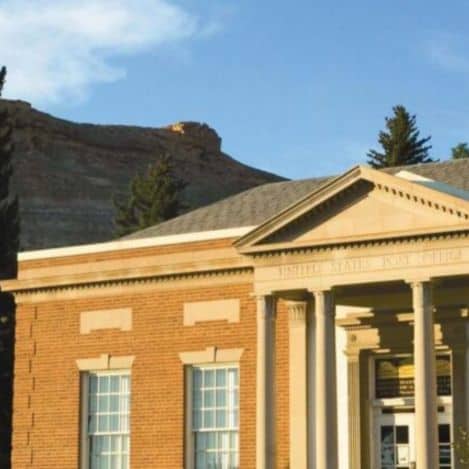 A brick post office building with white columns and a triangular pediment, set against a backdrop of rocky hills and blue sky.