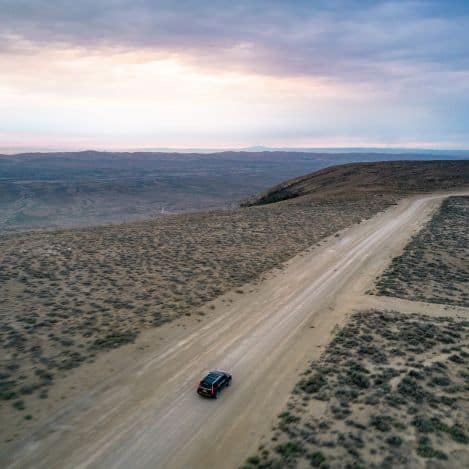 A black car drives along a dirt road through a barren, desert landscape under a cloudy sky at sunset.