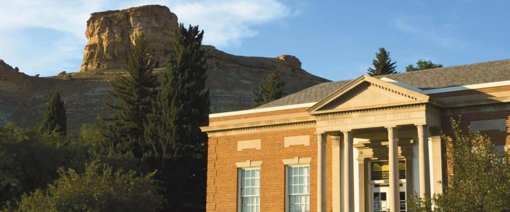 A brick building labeled United States Post Office stands in front of a rocky hill formation and tall trees under a clear sky.
