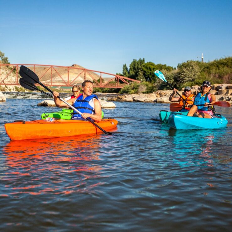 Three people kayaking on a river with life jackets; a red bridge and a pavilion are visible in the background under a clear sky.