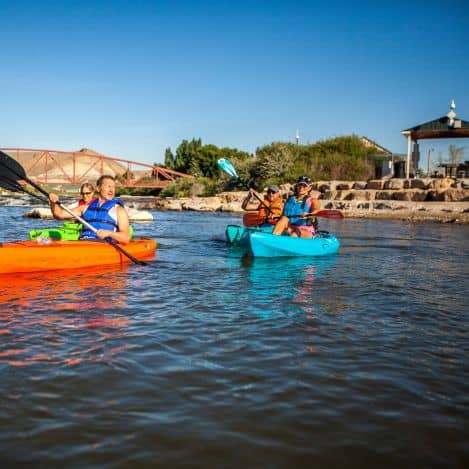 Four people wearing life jackets kayak on a calm river near a stone embankment, with a red bridge and a gazebo visible in the background under a clear blue sky.