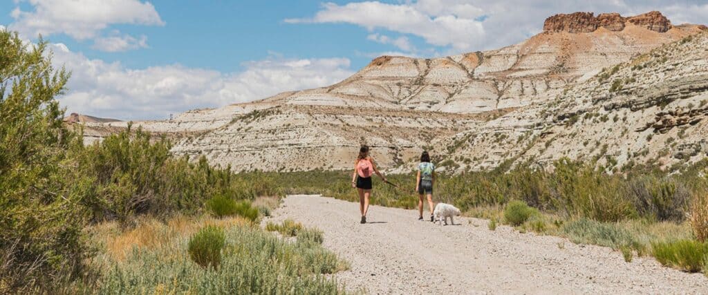Two people and a dog walk on a gravel trail through a dry, rocky landscape with layered hills under a partly cloudy sky.