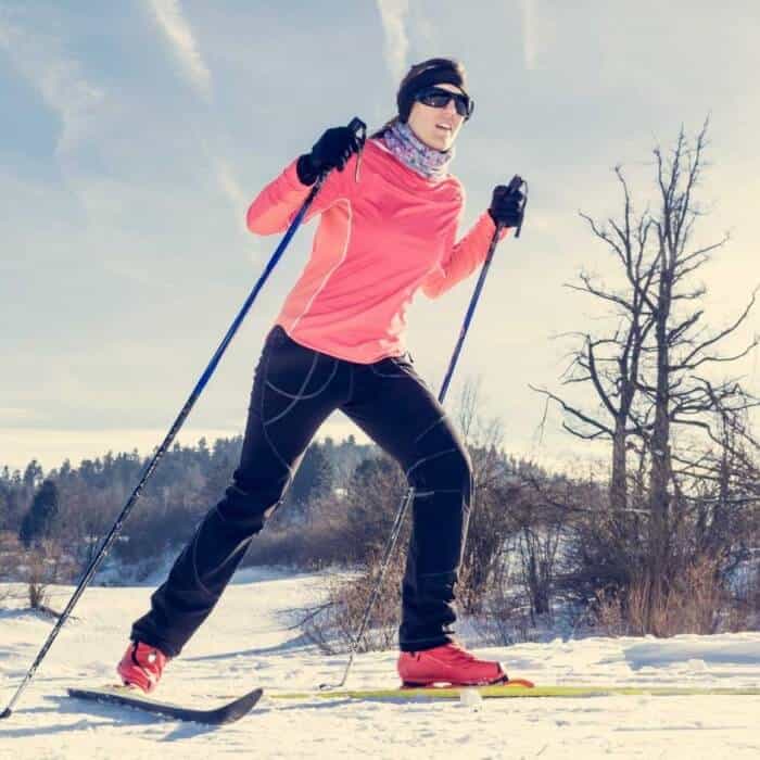 Person cross-country skiing on snowy ground, wearing a pink jacket, black pants, gloves, sunglasses, and a headband, with bare trees and a blue sky in the background.