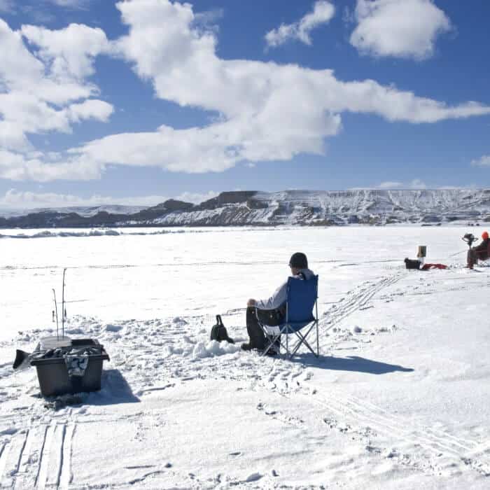 Two people are ice fishing on a snow-covered frozen lake, sitting on portable chairs with fishing gear nearby under a partly cloudy sky.