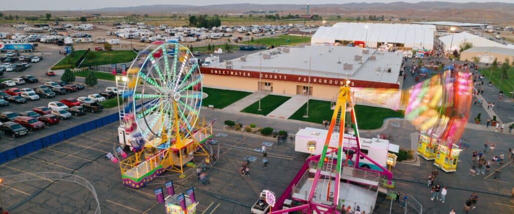 Aerial view of a fairground with a Ferris wheel, colorful rides, parked cars, and a building labeled "Sweetwater County Fairgrounds.