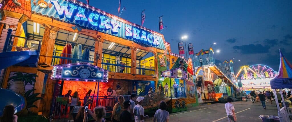 People walk past colorful, illuminated carnival attractions at dusk, including a building labeled "Wacky Shack" and various rides in the background.