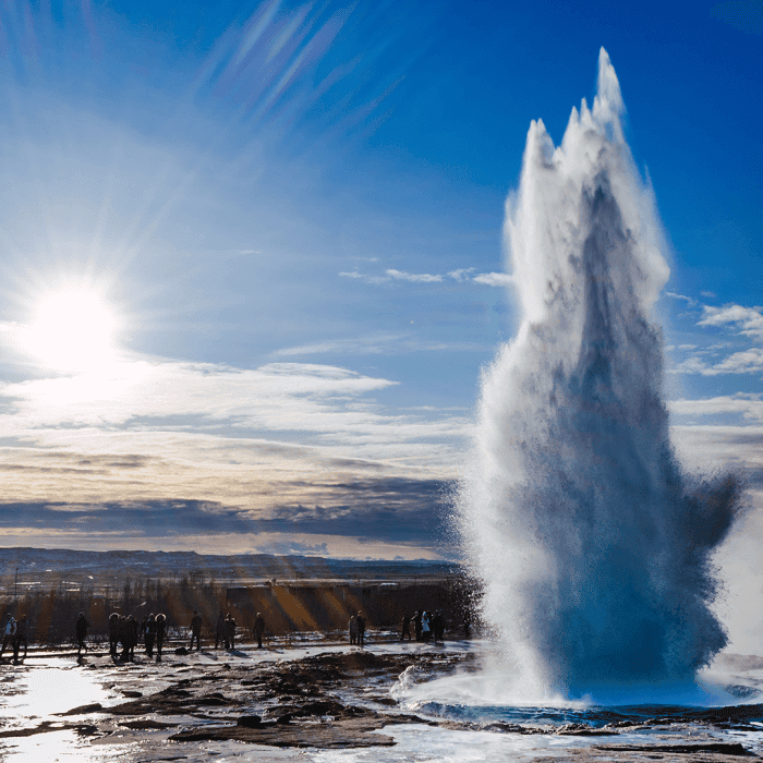A geyser erupts, shooting water high into the air under a bright sky, with people standing at a distance observing the scene.