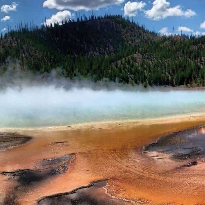 Colorful geothermal hot spring with orange, yellow, and blue hues, emitting steam, surrounded by forested hills and a partly cloudy sky.