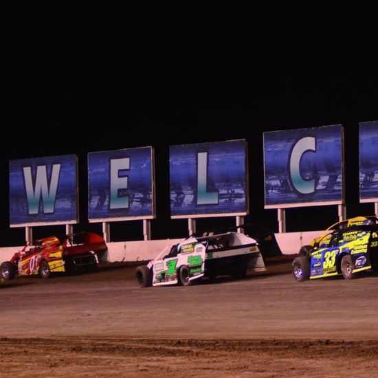 Four race cars drive on a dirt track at night in front of large signs spelling 
