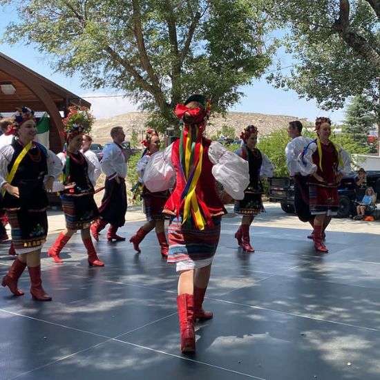 A group of people in traditional costumes perform a folk dance outdoors on a sunny day, surrounded by trees and spectators.