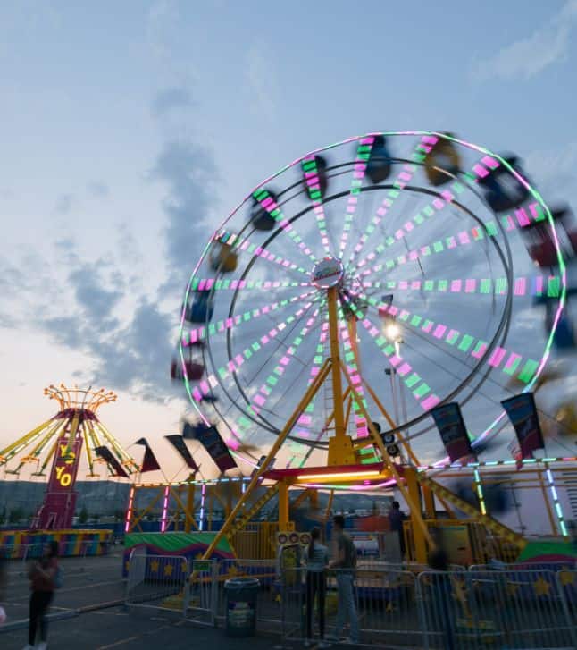 A Ferris wheel with colorful lights spins at dusk at a fairground, with another ride and people visible in the background.