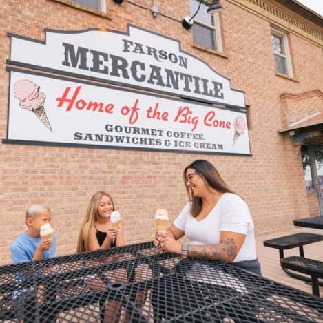 A woman and two children sit at an outdoor table eating ice cream cones in front of a sign that reads "Farson Mercantile, Home of the Big Cone.