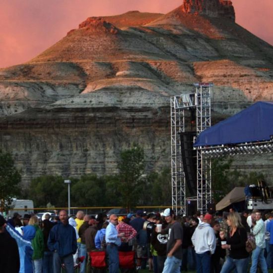A crowd gathers outdoors near a stage setup, with a rugged, layered hill and a pinkish sky in the background.