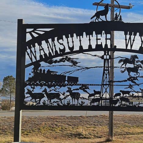 Metal sign reading "Wamsutter WY" features silhouettes of a wolf, train, windmill, horses, and wildlife against a partly cloudy sky background.