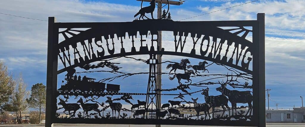 Metal sign reads "Wamsutter, Wyoming" with silhouettes of a train, horseback riders, wild animals, and trees against a blue sky background.