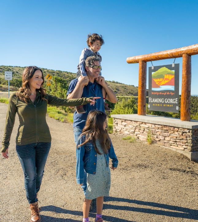 A family of four stands near a sign for Flaming Gorge Scenic Byway; the mother points ahead, the father carries a child on his shoulders, and another child stands in front.