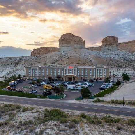 A hotel with a large parking lot is set against rocky cliffs at sunset, with several vehicles parked in front and a road in the foreground.
