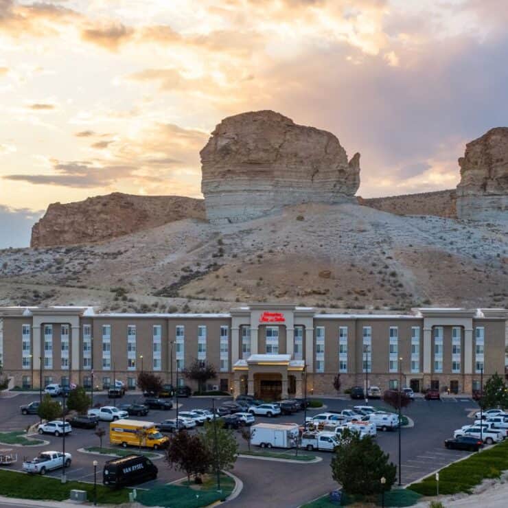 A large hotel stands in front of rocky hills at sunset, with numerous cars and trucks parked outside and dramatic clouds in the sky.