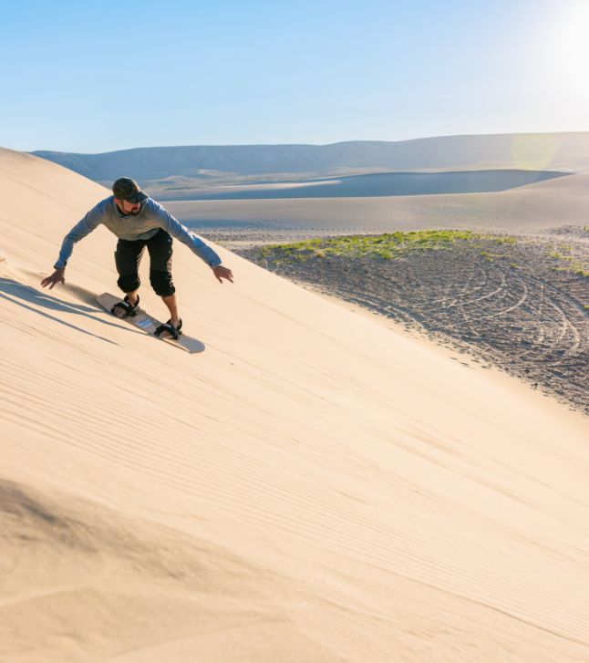 Person sandboarding down a sunlit sand dune in a desert landscape, wearing casual athletic clothing and sunglasses under a clear sky.