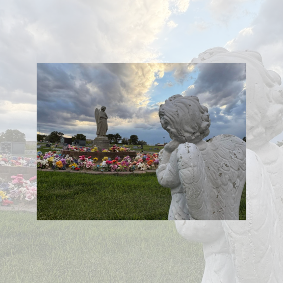 A white angel statue faces a grave adorned with colorful flowers under a cloudy sky in a cemetery.