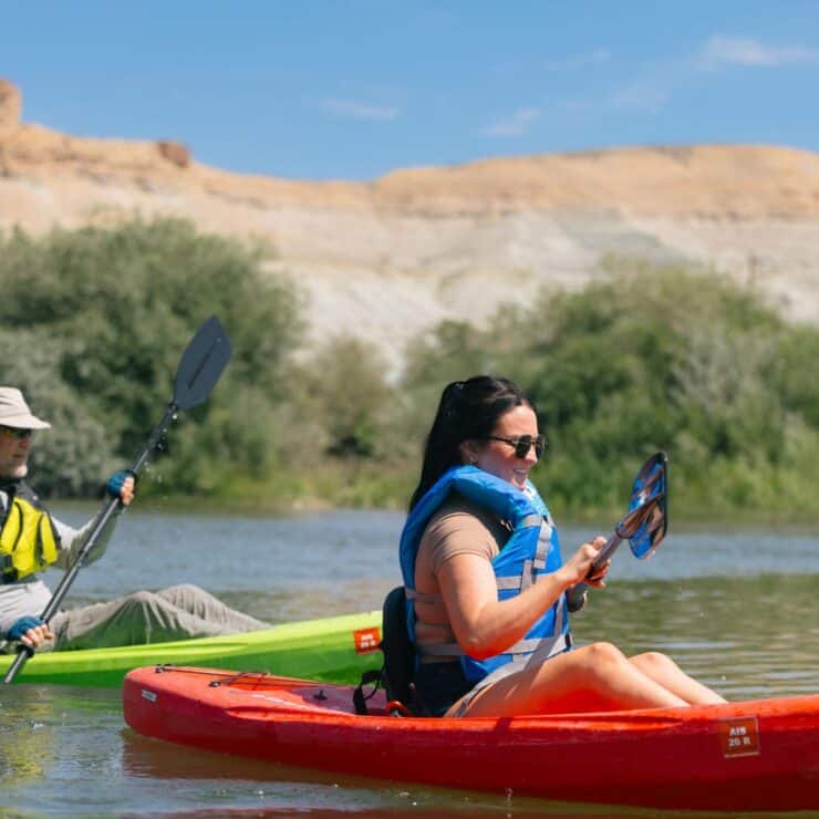 Two people paddle kayaks on a calm river with sandy cliffs and greenery in the background under a clear blue sky.