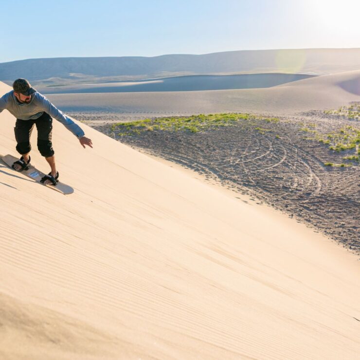 Person sandboarding down a large sandy dune under a clear sky, with desert landscape and distant hills in the background.